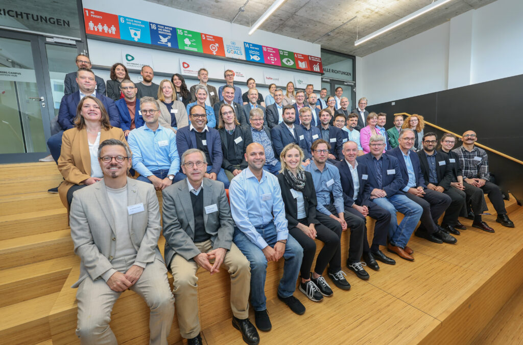 About 40-50 symposium participants sit in a tiered lecture hall and pose together for a group photo. // Etwa 40-50 Symposiumsteilnehmer sitzen in einem Stufenhörsaal und posieren zusammen für ein Gruppenfoto.